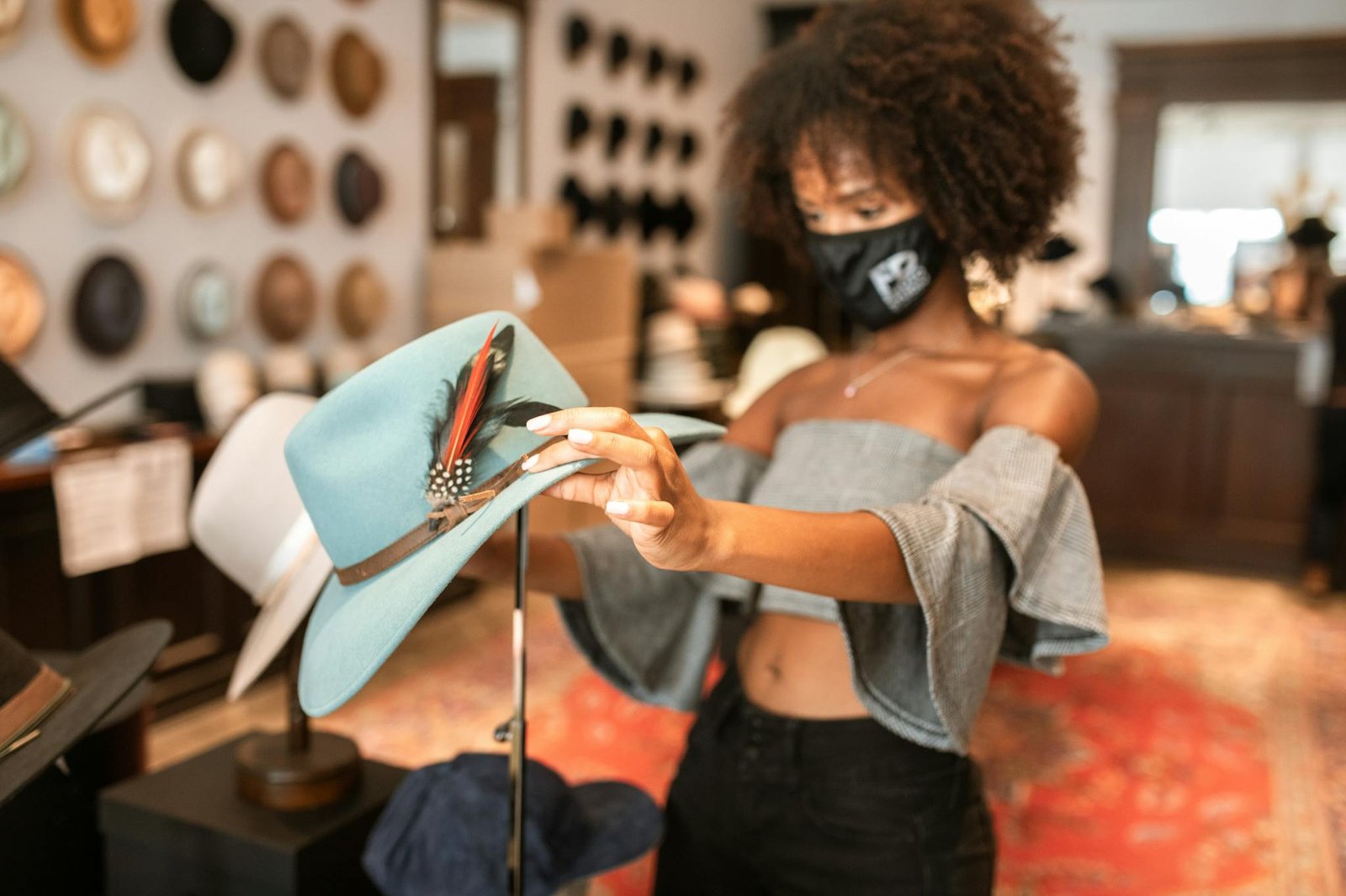 A fashionable woman with curly hair shops for hats in a stylish boutique, wearing a face mask and exploring various designs.